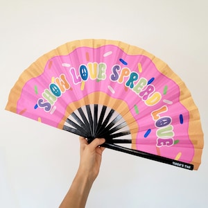 May include: Pink hand fan with a donut design and the text "Show Love Spread Love" in rainbow colors. The fan is made of paper and has a black wooden handle.