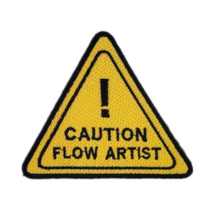 May include: Yellow triangular patch with a black border. The patch features a black exclamation mark above the words "CAUTION FLOW ARTIST" in black, all on a yellow background. The patch is likely an iron-on or sew-on patch.