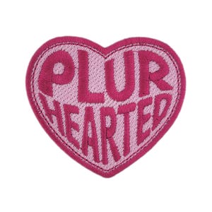 May include: A heart-shaped embroidered patch in shades of pink. The words "PLUR HEARTED" are stitched in a darker pink on a lighter pink background. The patch has a darker pink stitched border. The background is white.