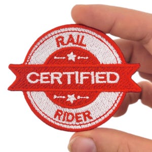 May include: A circular embroidered patch in red and white. The patch reads "RAIL CERTIFIED RIDER" in white text. The design includes star and line details above and below the central text.