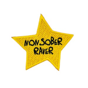 May include: A yellow star-shaped patch with the embroidered words "NON SOBER RAVER" in black. The patch has a stitched border and is designed to be attached to clothing or accessories.