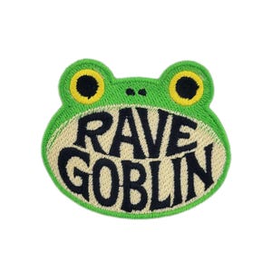 May include: Embroidered patch in the shape of a green frog with yellow eyes. The patch features the words "RAVE GOBLIN" in black, stylized lettering on a beige background. The patch is designed for clothing or accessories.