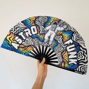 May include: A black and white hand fan with a rainbow swirl pattern. The fan features an astronaut in a space suit and the text "Astro Funk".