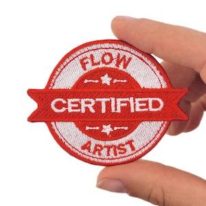 May include: A circular embroidered patch in red and white. The patch reads "FLOW CERTIFIED ARTIST" in white lettering on a red banner and border. The design also includes two small stars. The patch is held in a hand.