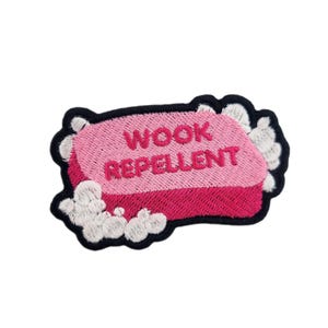 May include: Embroidered patch with a pink soap bar design and the words "WOOK REPELLENT" in pink. It has a black border and white suds around the edges. Ideal for customising clothing or accessories, such as jackets, bags, or other fabric items.