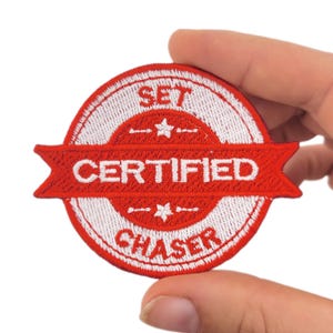 May include: A red and white embroidered patch with the words "SET CERTIFIED CHASER". The patch is circular with a banner across the middle. The patch is held by a hand.