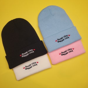 May include: Two reversible knit beanies. One beanie is black with white trim and the other is light blue with pink trim. Both beanies have the text "Show Love Spread Love" embroidered on them.