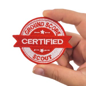 May include: A circular, red and white embroidered patch featuring the text "GROUND SCORE CERTIFIED SCOUT" in white. The patch is held in a hand. The design includes a banner shape and star details.
