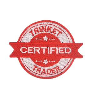 Puede incluir: Un parche bordado rojo y blanco con las palabras "TRINKET CERTIFIED TRADER". El parche es circular con una pancarta en el centro. También se incluyen estrellas y diseños florales.