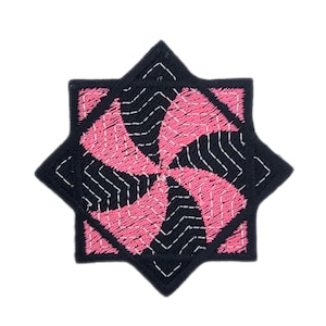 May include: Embroidered patch in a starburst design. The patch features a black eight-pointed star shape with a pink and black spiral pattern in the centre. White stitching outlines the design.