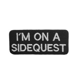 May include: Black rectangular patch with white embroidered text reading "I'M ON A SIDEQUEST". The patch has rounded corners and is set against a white background. The text is centered.