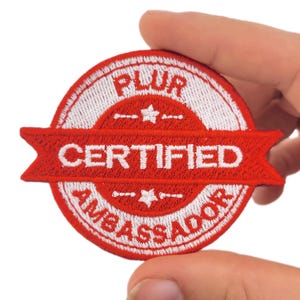 May include: An embroidered patch with a red and white design. The patch is circular with a red banner across the middle, displaying the words "CERTIFIED AMBASSADOR". The word "PLUR" is at the top.