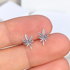 May include: A pair of small, silver-colored stud earrings shaped like cannabis leaves. The earrings are detailed with veining and are displayed on a light background.