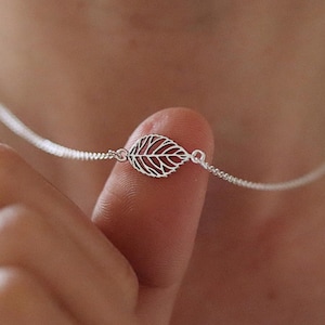 May include: A delicate silver necklace featuring a leaf pendant. The leaf design is intricately detailed with openwork, showcasing the veins of the leaf. The necklace is shown against a blurred background, highlighting the jewelry.