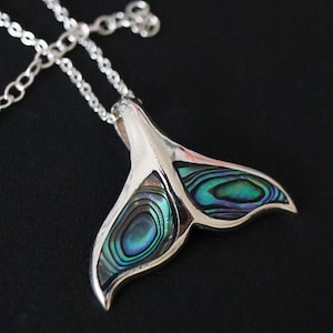 May include: A silver chain necklace with a pendant shaped like a whale tail. The tail is made of iridescent abalone shell with green, blue, and purple hues.