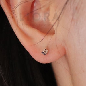 May include: A close-up of a person's ear with a small silver dice earring. The earring is a simple design with black dots on each side.