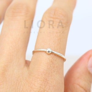 May include: A simple silver ring with a small bead in the centre. The ring is on a person's finger.