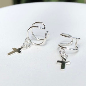 Może przedstawiać: Para srebrnych kolczyków na ucho z wiszącym krzyżykiem. Kolczyki wykonane są z cienkiego, skręconego drutu i są przeznaczone do noszenia na górnej części ucha.