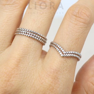 May include: Two silver rings on a finger. One ring is a simple band with three rows of small beads. The other ring is a V-shaped band with two rows of small beads.