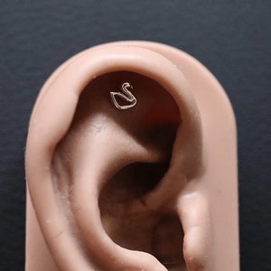 May include: A close-up of a faux ear with a small silver swan earring in the cartilage piercing.