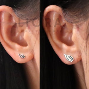 May include: A pair of silver wing earrings. The earrings are small and delicate, and they are shown on a model's ear.