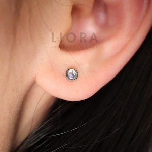 Puede incluir: Primer plano de una oreja con un pequeño pendiente de plata con una piedra de ópalo redonda e iridiscente.