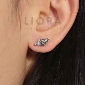 Puede incluir: Un pendiente de concha plateada se lleva en una oreja. El pendiente es una réplica en miniatura detallada de una concha. La joya forma parte de la colección Liora Jewelry House.