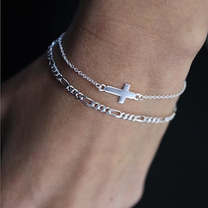 May include: Two silver bracelets on a wrist. The top bracelet has a small silver cross charm. The bottom bracelet is a thicker chain.