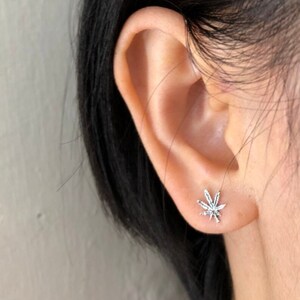 May include: A silver-colored stud earring in the shape of a cannabis leaf is shown on an ear. The earring has detailed veining and a shiny finish. The ear is in focus against a blurred background.