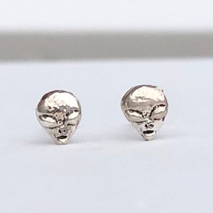 Può includere: Un paio di orecchini a perno in argento a forma di testa aliena. Gli orecchini sono piccoli e dettagliati, con gli occhi, il naso e la bocca dell'alieno chiaramente visibili.