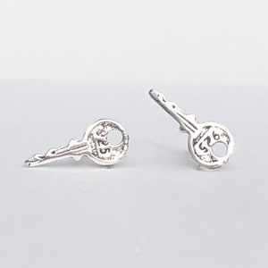 Puede incluir: Un par de pendientes de plata en forma de llave. Cada pendiente tiene una pequeña llave con una cabeza redonda y el número "526" grabado en ella.