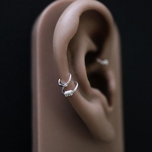 May include: Two silver hoop earrings, one with a simple circular design and the other with a coiled wire design, are worn in the cartilage of a model's ear.