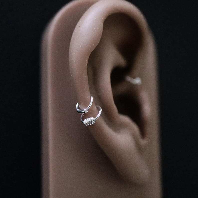 May include: Two silver hoop earrings, one with a simple circular design and the other with a coiled wire design, are worn in the cartilage of a model's ear.