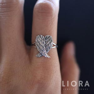 Op de afbeelding: Zilveren ring met een hartvormig engelenvleugelontwerp. De vleugels zijn gedetailleerd met individuele veren. De ring wordt op een vinger getoond. Het logo van het sieradenhuis "LIORA" is zichtbaar in de afbeelding.