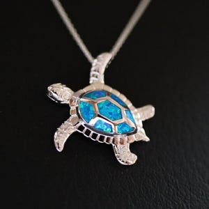 Puede incluir: Un collar con un colgante de tortuga de plata con una incrustación de ópalo azul. La tortuga está detallada con líneas grabadas y el ópalo es de un color azul vibrante.
