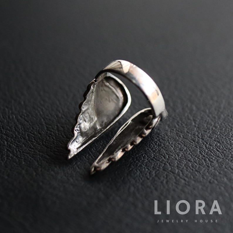 May include: A silver-toned ring with a wing design. The ring features two detailed wing-shaped elements extending from the band. The wings have textured surfaces and are positioned to create a unique, open design. The ring is set against a dark background.