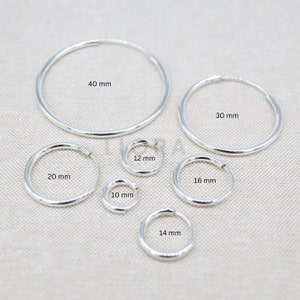 Puede incluir: Un conjunto de pendientes de aro de plata en varios tama&ntilde;os, que van desde los 10 mm hasta los 40 mm de di&aacute;metro. Los pendientes est&aacute;n dispuestos en semic&iacute;rculo sobre una superficie texturizada.