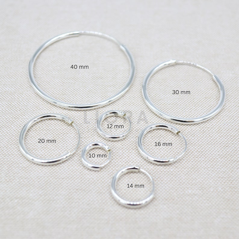 Puede incluir: Un conjunto de pendientes de aro de plata en varios tama&ntilde;os, que van desde los 10 mm hasta los 40 mm de di&aacute;metro. Los pendientes est&aacute;n dispuestos en semic&iacute;rculo sobre una superficie texturizada.