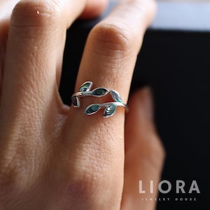 May include: A silver ring with a leaf design. The ring features two curving bands that meet at the top, each adorned with three leaf-shaped inlays with a green and iridescent finish. The ring is shown on a finger.