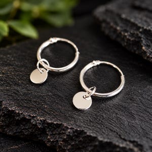 May include: A pair of silver hoop earrings, each featuring a small, smooth, circular disc charm suspended from the hoop. The earrings have a polished, reflective metallic finish and a simple, circular design with a hinged closure mechanism.