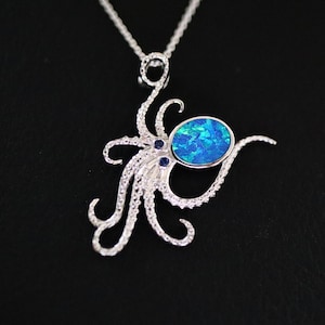 Puede incluir: Un colgante de plata en forma de pulpo con una piedra preciosa de ópalo azul y dos piedras azules más pequeñas para los ojos. El colgante está en una cadena de plata.