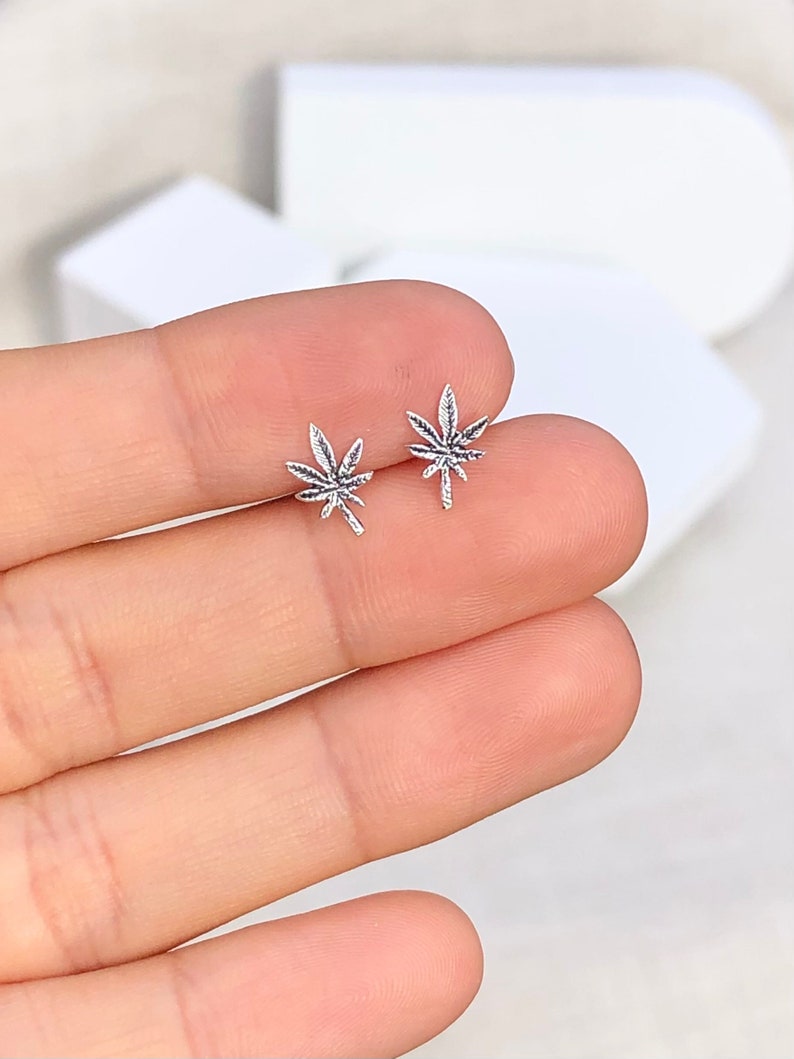 May include: A pair of small, silver-colored stud earrings shaped like cannabis leaves. The earrings are detailed with veining and are displayed on a light background.