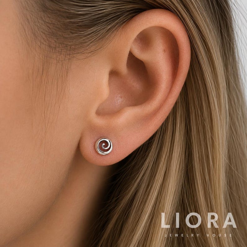 Op de afbeelding: Een kleine, zilverkleurige oorsteker met een cirkelvormig spiraalontwerp. De oorsteker is geplaatst op de oorlel. De tekst LIORA JEWELRY HOUSE is zichtbaar in de rechterbenedenhoek van de afbeelding, geschreven in een strak, schreefloos lettertype.