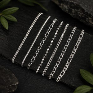 Puede incluir: Una colección de seis cadenas de metal plateado dispuestas en paralelo. El conjunto incluye una cadena de bordillo fina, una cadena de serpiente lisa, una cadena de eslabones ovalados alargados, una cadena de cuentas, una cadena de cable y una cadena figaro.