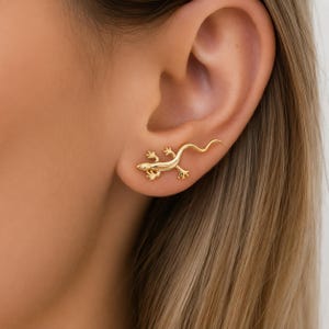 Puede incluir: Un pendiente dorado con forma de lagartija, que presenta un cuerpo detallado con cuatro patas y una cola curva. El pendiente está colocado horizontalmente a lo largo del lóbulo, con la cabeza de la lagartija hacia adelante y la cola hacia atrás.