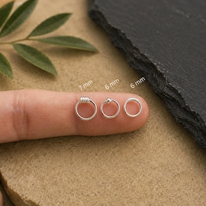 Puede incluir: Tres aros de plata con diseños variados. El aro de la izquierda presenta un detalle de alambre enrollado y está marcado con 7 mm. El aro central tiene una pequeña cuenta esférica y está marcado con 6 mm. El aro de la derecha es liso y marca 6 mm.