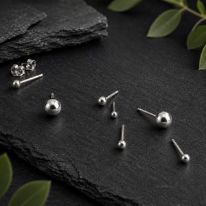 May include: A collection of silver-toned ball stud earrings in various sizes, each featuring a smooth, polished spherical head attached to a straight post, accompanied by small metal earring backs for secure fastening to the earlobe.