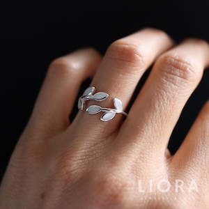 May include: A silver ring with a leaf design. The ring has two sets of leaves, each with three leaves, that wrap around the finger. The ring is shown on a person's finger. The brand name "LIORA JEWELRY HOUSE" is visible in the image.