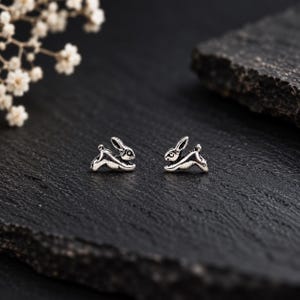 May include: A pair of silver-toned stud earrings shaped like small, leaping rabbits. The earrings feature a detailed, oxidized finish that highlights the ears, body, and tail of the rabbit figures, creating a distinct contrast against the metal surface.