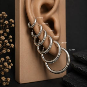 May include: A set of six polished silver hoop earrings arranged in increasing size from 10 mm to 20 mm. Each hoop is labelled with its corresponding diameter in millimetres, showcasing a graduated collection of circular metal jewellery pieces.